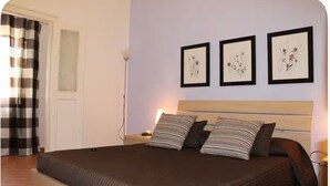 Cama bem arrumada com almofadas listradas, mesa de cabeceira com abajur e três quadros florais na parede.