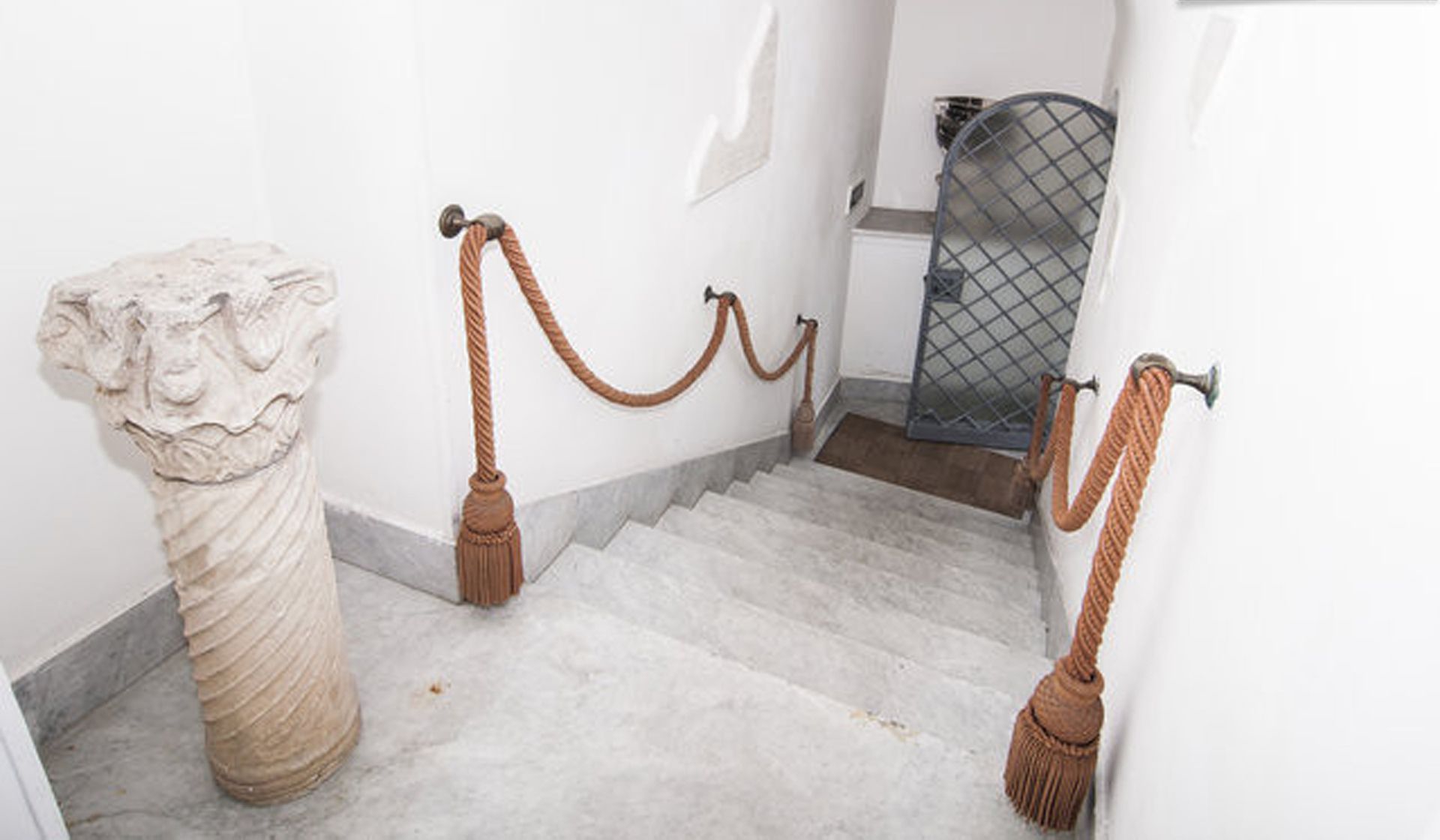 Una escalera de mármol con una barandilla de cuerda, una pared blanca y una reja metálica al fondo.
