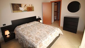 A bedroom with a bed, a nightstand with a lamp, a dresser, and a door leading to another room. - Geranei Appartamento Bilocale a 50 metri dal mare (TAGGIA)