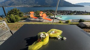 Un espace aménagé au bord de la piscine, avec des chaises longues, une table ornée d’un filet jaune et de fleurs, et une vue sur les montagnes et un lac.