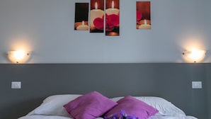 Un lit bien fait, avec du linge de lit blanc et deux coussins violets, un luminaire fixé au mur et une série de photographies encadrées représentant des bougies et des fleurs.
