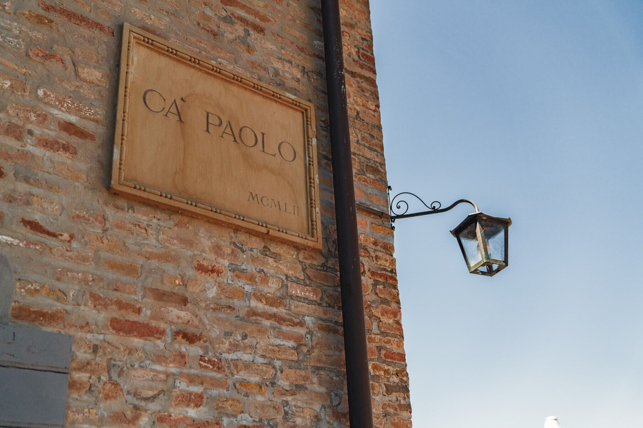 Un letrero de madera con la inscripción "CA PAOLO" y "MCMLII" sobre una pared de ladrillo, un poste de luz metálico y un cielo despejado.
