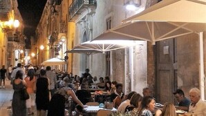 Une rue étroite bordée d’immeubles, avec des terrasses de cafés et des gens qui déjeunent ou dînent en plein air.
