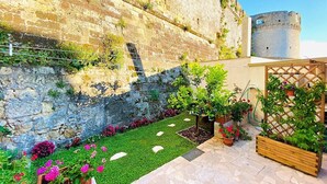 Une cour avec un mur en pierre, un petit jardin avec du gazon synthétique et des plantes en pot, et une clôture en bois.