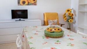 A dining table with a floral tablecloth, a green bowl with a sunflower design, a television on a white cabinet, a yellow chair with a floral cushion, and a sunflower vase on a white shelf.