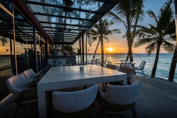 An outdoor dining area with a view of the sunset over the ocean, surrounded by palm trees.