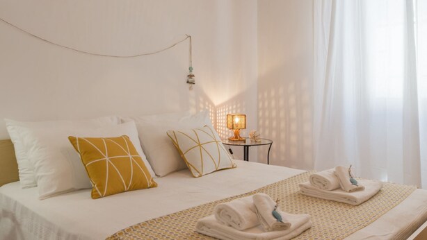 Ein Hotelzimmer mit einem ordentlich bezogenen Bett, einem Nachttisch mit Lampe und einem Fenster mit durchsichtigen Vorhängen.