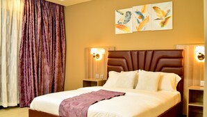 Uma cama bem arrumada com cabeceira marrom, duas mesas de cabeceira com abajures e uma janela grande com cortinas.