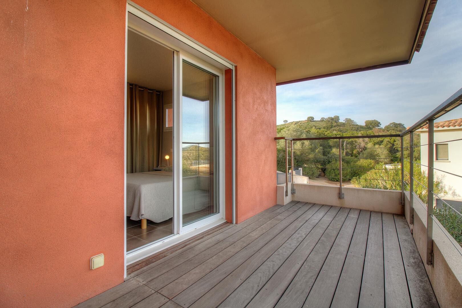 Ein Balkon mit Holzboden, eine Schiebetür zu einem Schlafzimmer und Blick auf eine hügelige Landschaft.