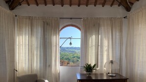 Une pièce confortable avec un canapé, un bureau et une vue sur l’extérieur, visible à travers une fenêtre ornée de rideaux.