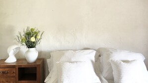 Une chambre à coucher avec du linge de lit blanc, une table de chevet en bois ornée d’un vase de fleurs et un mur blanc.
