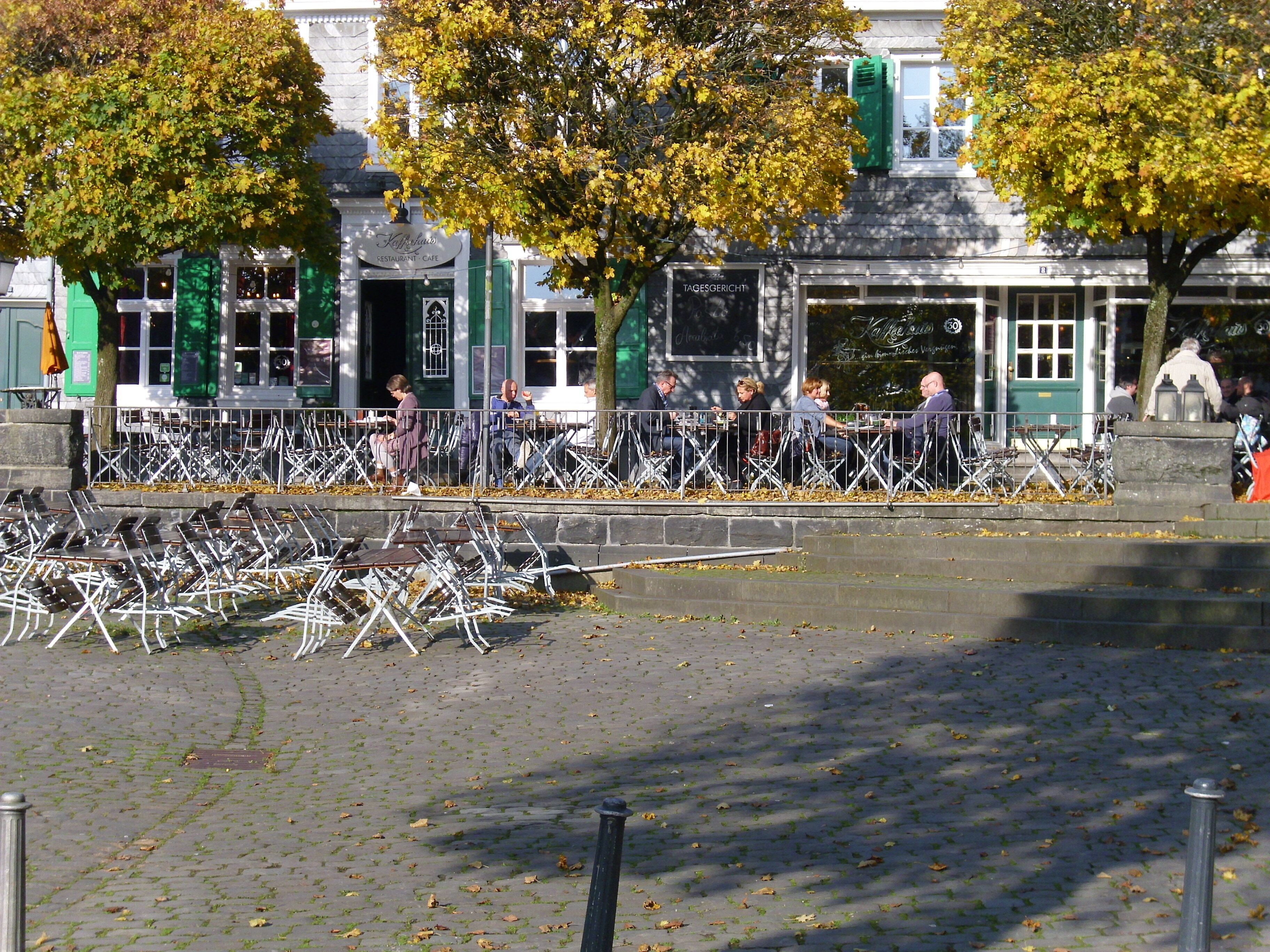 Een café met terras, een stenen muur en bomen met herfstbladeren.