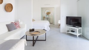 Een moderne, minimalistisch ingerichte hotelkamer met een witte bank, een ronde salontafel, een bed met witte lakens, een televisie op een witte voet en een badkamer in de achtergrond.