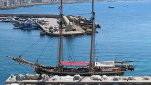 Ein Hafen mit einem großen Segelschiff, das anlegt, umgeben von kleineren Booten und einer Stadtkulisse im Hintergrund.