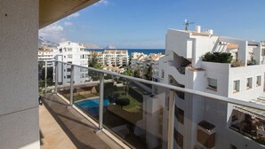 Un balcon doté d’une balustrade en verre, offrant une vue sur une piscine et un quartier résidentiel avec des bâtiments blancs et un ciel d’un bleu éclatant.