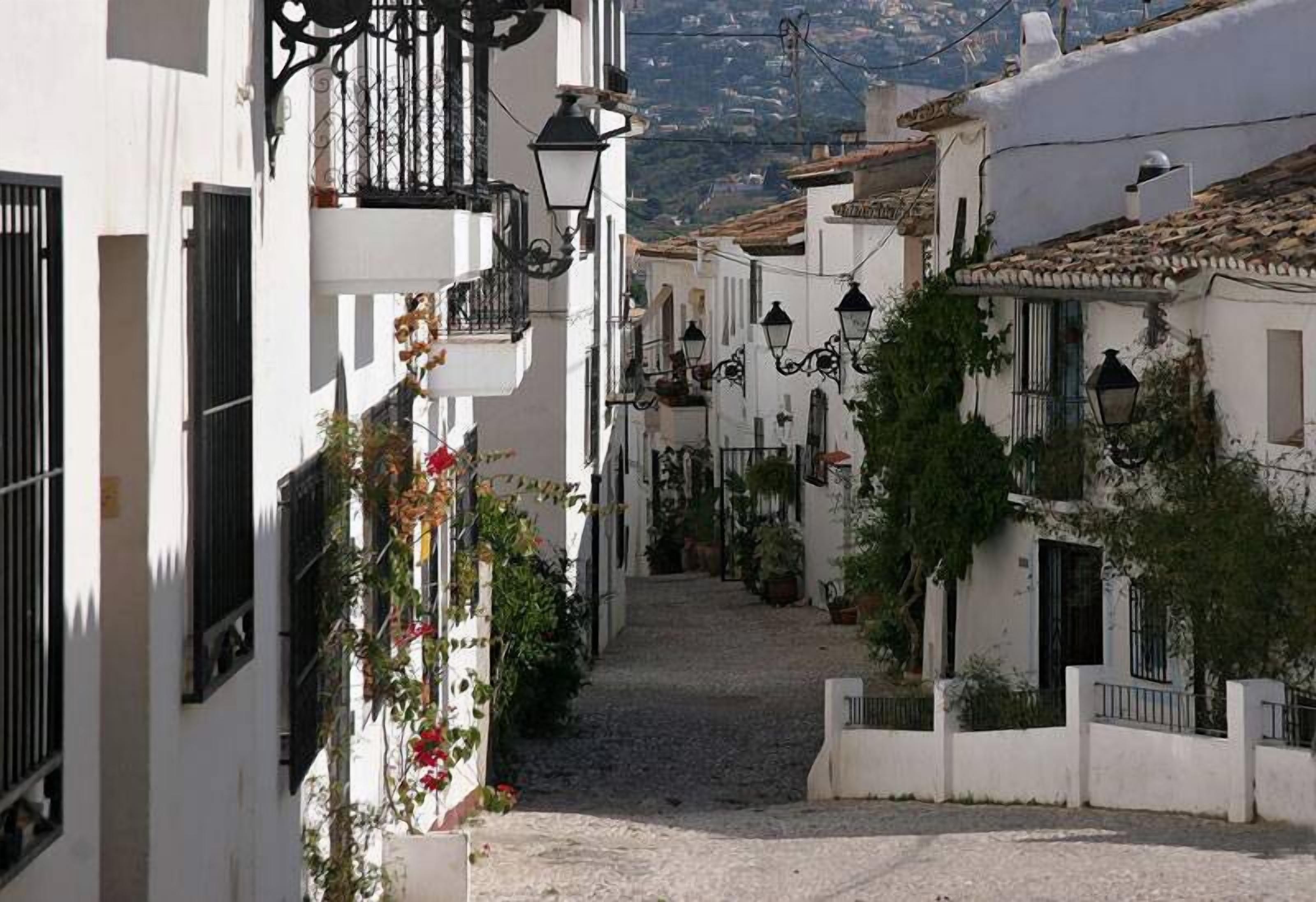 Une ruelle pavée étroite, bordée de bâtiments blancs aux volets noirs et aux balcons en fer forgé.