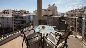 Un balcon avec une table et des chaises en verre, donnant sur un paysage urbain avec des immeubles et un ciel dégagé.