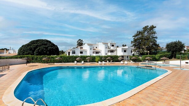 Uma piscina com um deck ladrilhado, rodeada por espreguiçadeiras e guarda-sóis, com um edifício ao fundo.
