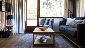 Una sala de estar acogedora con piso de madera, un sofá, una mesa de centro con libros, un televisor y un ventanal con vista a árboles.