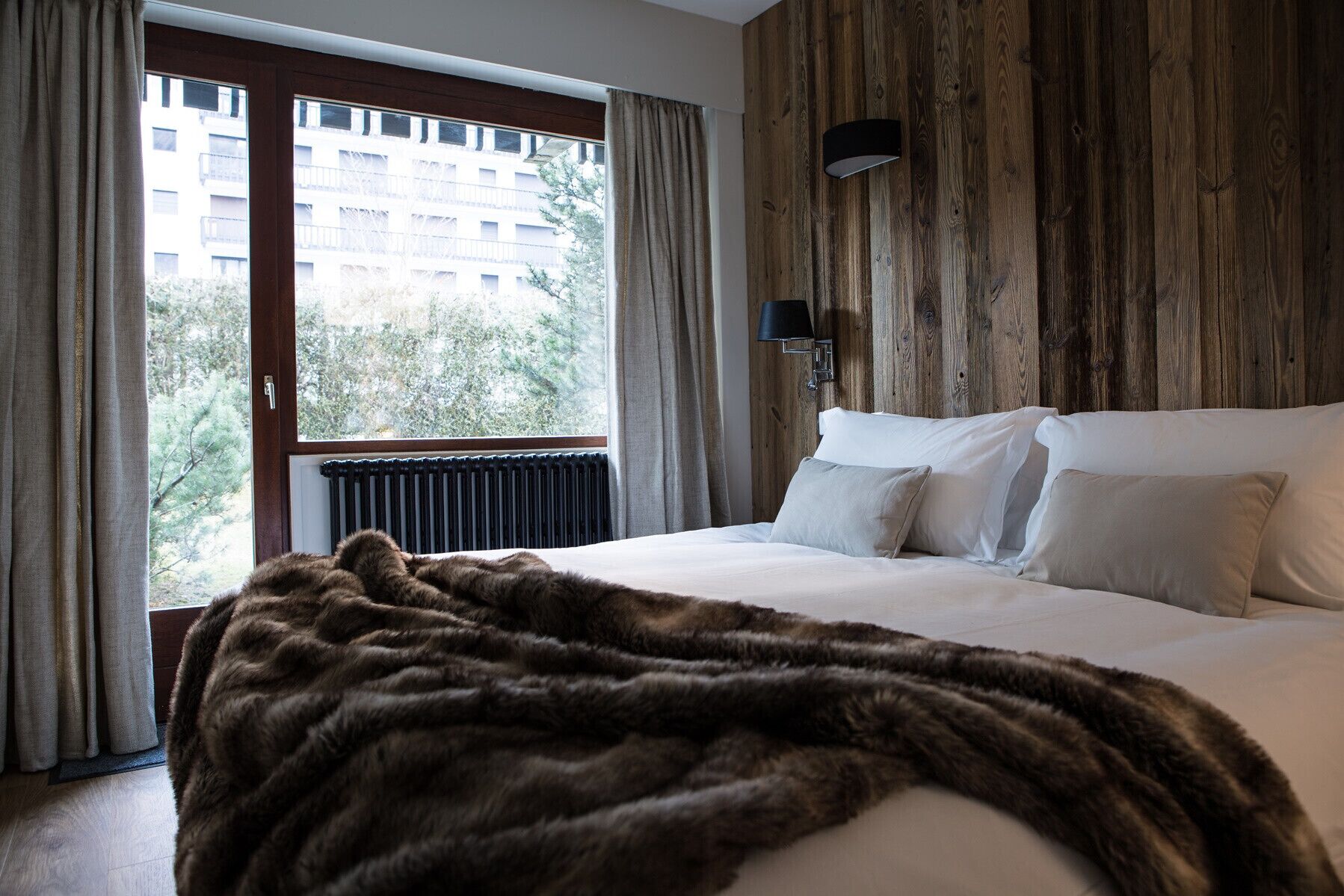 A bedroom with a large bed, a wooden wall panel, a window with curtains, and a radiator.