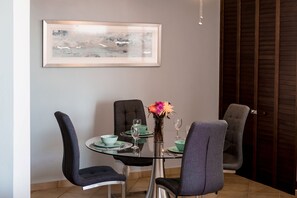 A dining area with a round glass table, four grey chairs, a vase with flowers, and a framed abstract painting on the wall.