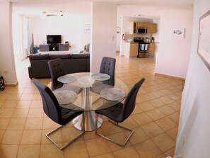 A modern living room with a dark grey sofa, a glass-top dining table with four chairs, and a kitchen in the background.
