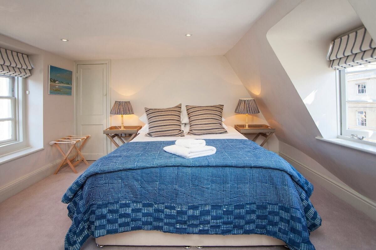 Une chambre à coucher avec un lit, deux lampes de chevet, une fenêtre avec des rideaux, un tableau au mur et une petite table avec une lampe.