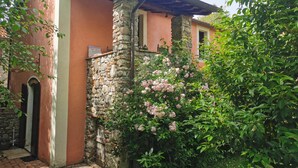 A rustic building with a stone wall and a wooden door, surrounded by lush greenery and pink flowers.