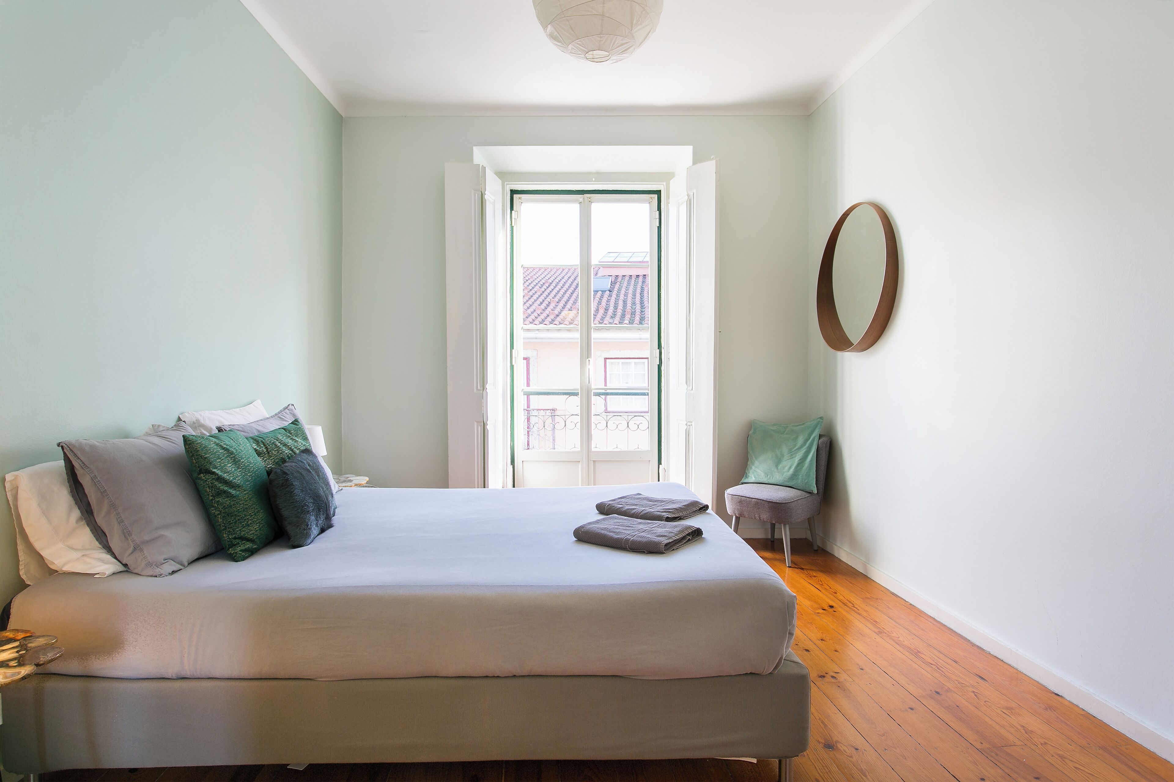 A bedroom with a bed, pillows, a small stool, and a window with a view of buildings.