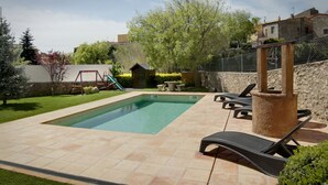Una piscina rectangular con solárium de baldosas, rodeada por un muro de piedra y una zona ajardinada. Se observan sillones de descanso y una pequeña construcción de madera al fondo.