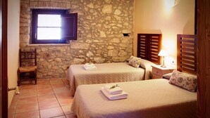 Una habitación con dos camas, una pared de piedra, una ventana, una silla y una mesita de noche con una lámpara.