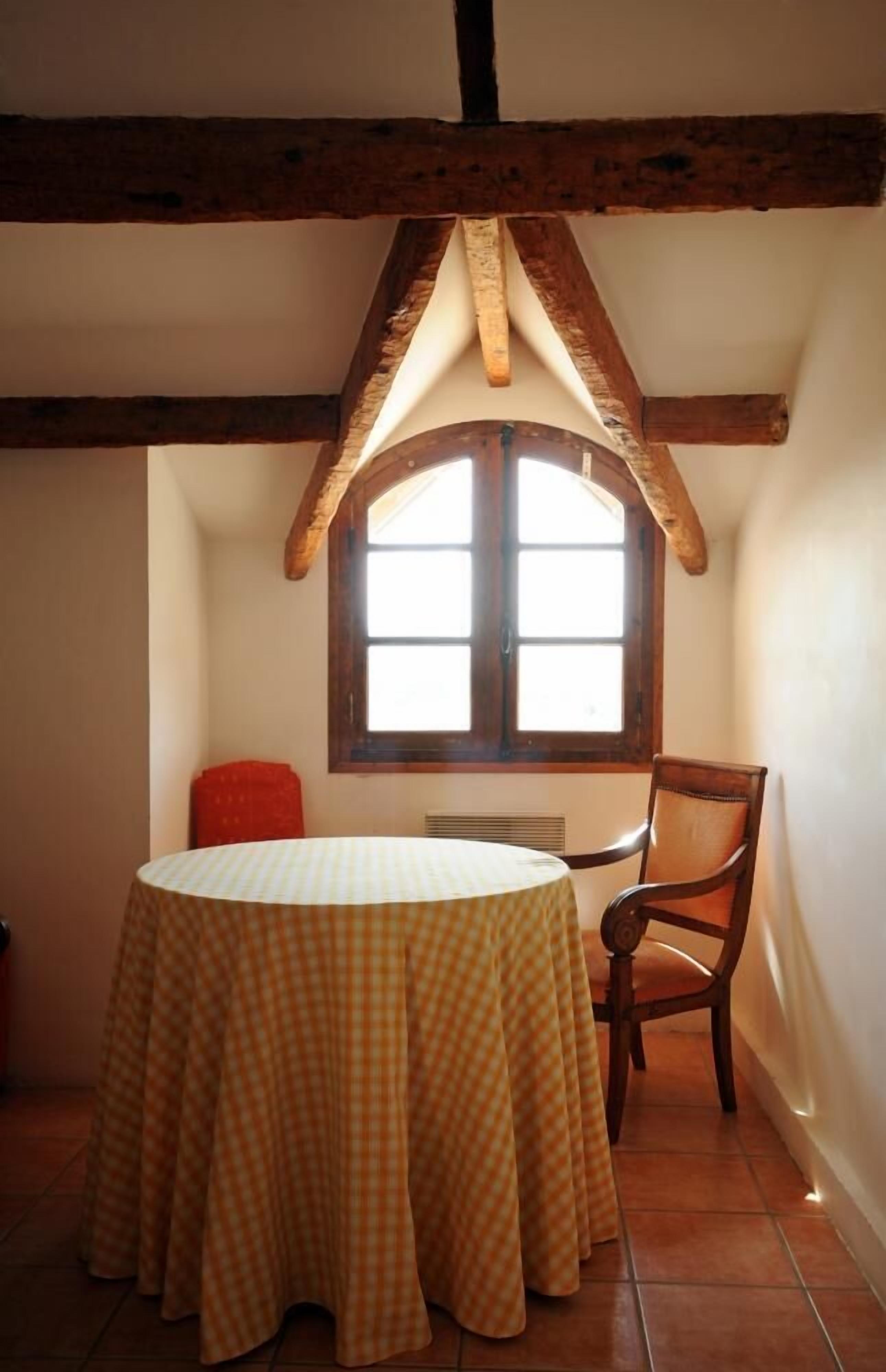 A dining area with a round table covered by a checkered tablecloth, a wooden chair, and a window with wooden shutters.