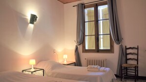 Une chambre avec deux lits, une fenêtre avec des rideaux, un radiateur, une chaise et une lampe fixée au mur.