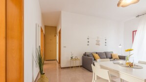 A modern living room with a grey sofa, white dining table, and chairs. There is a small side table with a plant and a lamp. - Holiday house in Torre Lapillo beach (TL23) (Torre Lapillo)