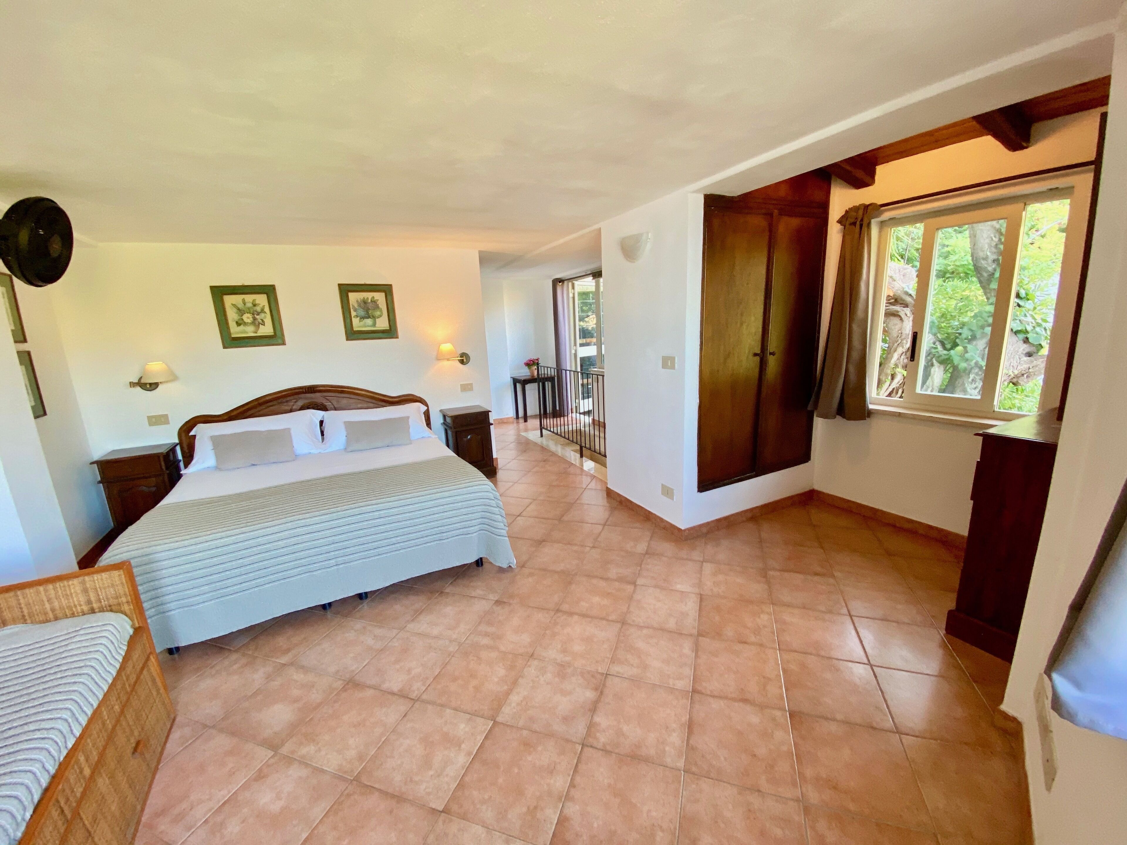 A bedroom with a large bed, a small bedside table, a nightstand, a window with curtains, and a door leading to another room.