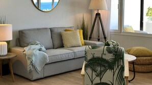Een woonkamer met een grijze bank, een ronde spiegel, een vloerlamp en uitzicht op de zee door een raam.