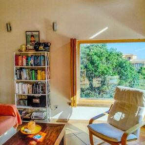 Um quarto com uma grande janela, uma estante de livros, uma cadeira e uma mesa com uma vela.