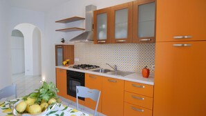 A kitchen with orange cabinets, a gas stove, and a sink. There is a table with a lemon-patterned tablecloth and a chair. - CONCA "Marechiaro" AMALFI COAST (Conca dei Marini)