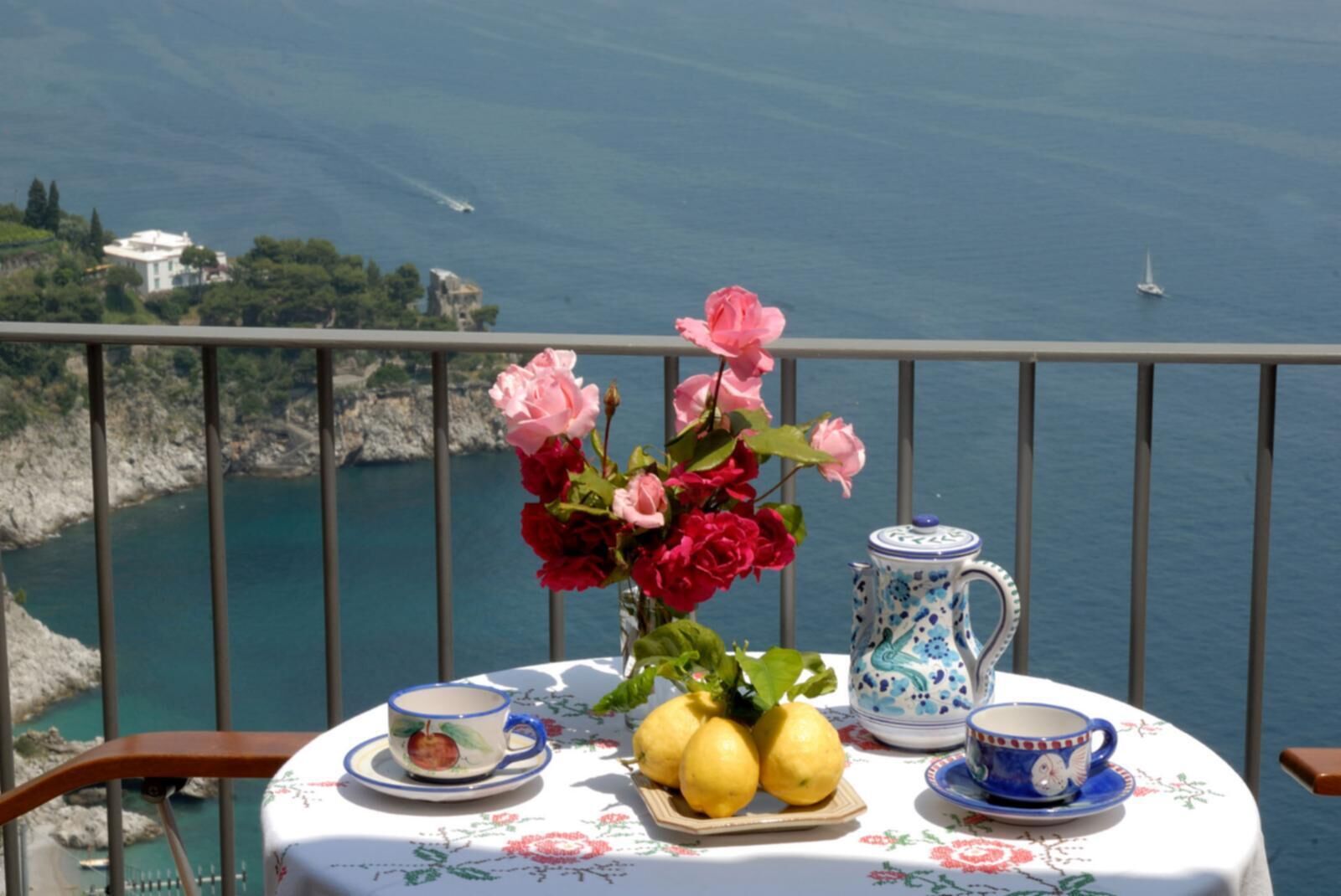 Uma mesa com toalha florida, um vaso de flores, limões, um bule e xícaras, com uma paisagem costeira ao fundo.