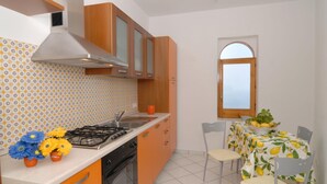 A compact kitchen with a gas stove, sink, and orange cabinets. There is a dining area with a table set for two and a window with a view. - CONCA "Marechiaro" AMALFI COAST (Conca dei Marini)