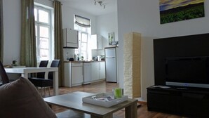 Ein modernes Wohnzimmer mit Flachbildfernseher, eine Essecke mit Tisch und Stühlen, eine Küche mit weißen Schränken und einem Kühlschrank sowie ein großes Fenster mit Vorhängen.