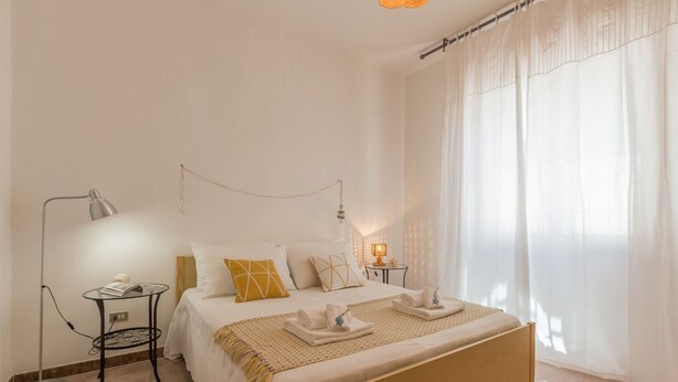 Une chambre à coucher bien rangée, avec un lit, des tables de chevet, une lampe et une fenêtre ornée de rideaux légers.