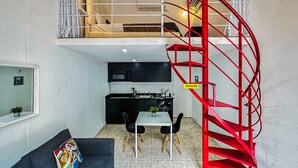 A compact living space with a kitchenette, a sofa with a colorful pillow, a dining area, and a red spiral staircase. - Loft Aparment Steps From Nightlife, Gladiolas II /Loft + Room /Free WIFI - 2101 (Ponce Pueblo)