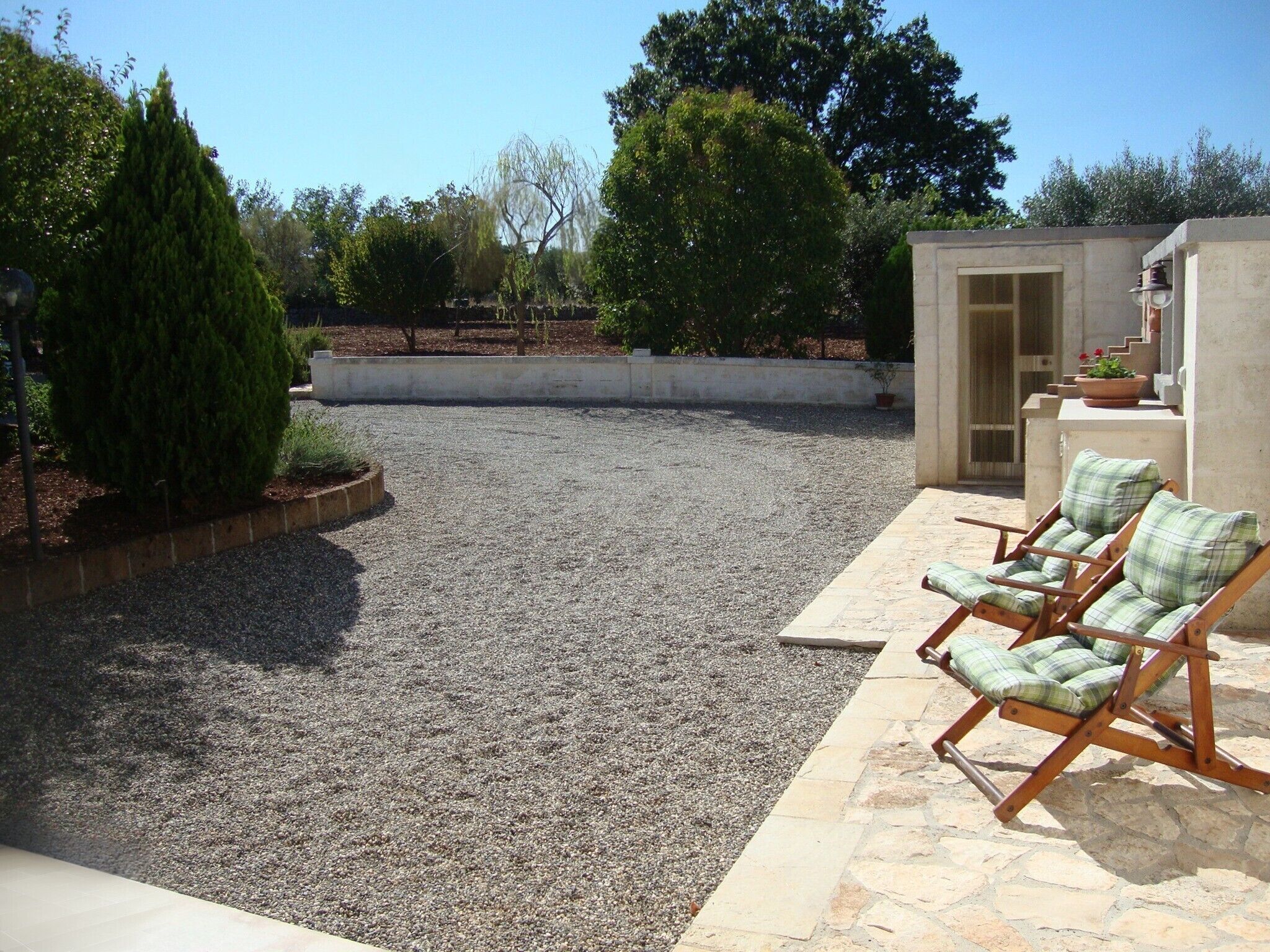 A patio area with a gravel surface, two wooden lounge chairs with green cushions, a small concrete structure with a door, and a potted plant with red flowers.