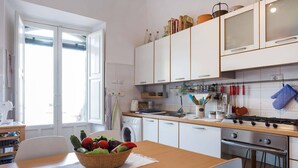 Une cuisine avec des meubles blancs, une table en bois sur laquelle se trouve un panier de fruits, un lave-linge et une cuisinière.