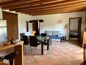 A dining area with a glass table and chairs, a wooden ceiling, and a sofa in the background.