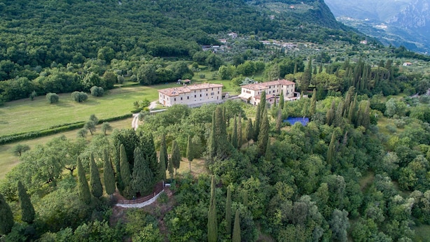 An aerial view of a rural estate with two large buildings surrounded by dense greenery and a mountainous backdrop. - Cozy Apartment for Two on the Hills of Lake Garda (Toscolano Maderno)