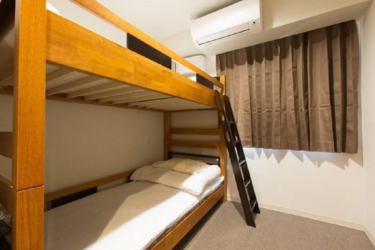Een stapelbed met houten frame, matras en kussen. Een ladder is bevestigd aan het bed. Een airconditioning is aan de muur boven het bed gemonteerd.