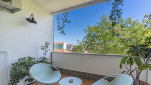 En balkon med to stole, et lille bord, en potteplante og udsigt til bygninger og træer.