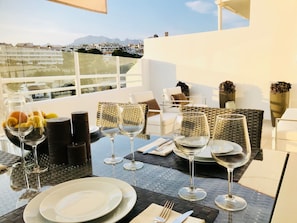 Una mesa de comedor dispuesta con copas de vino, platos y un bol de frutas en un balcón con vistas a la ciudad.